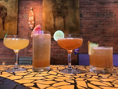Four colorful cocktails on a patterned table, featuring garnishes like lime, watermelon, and pineapple.