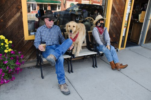 A man in a cowboy hat sits on a bench with a golden retriever, while a mannequin in a cowboy outfit sits beside them.