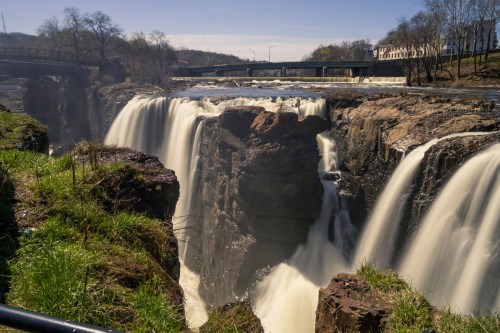 A scenic view of a waterfall cascading over rocky cliffs, surrounded by greenery and a bridge in the background.