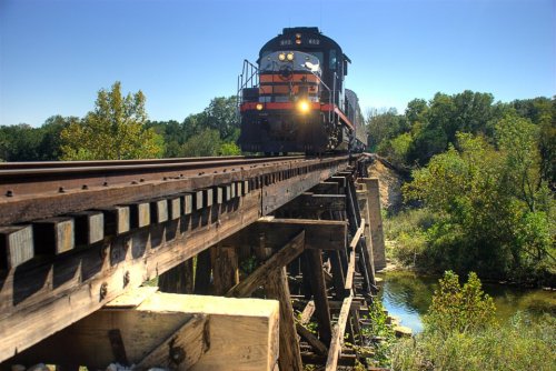 A train crosses a wooden trestle bridge over a river, surrounded by lush greenery under a clear blue sky.