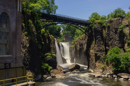 A scenic view of a waterfall cascading between rocky cliffs, with a bridge overhead and lush greenery surrounding the area.
