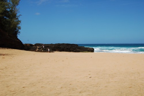 A sandy beach with gentle waves, rocky formations, and a clear blue sky in the background.