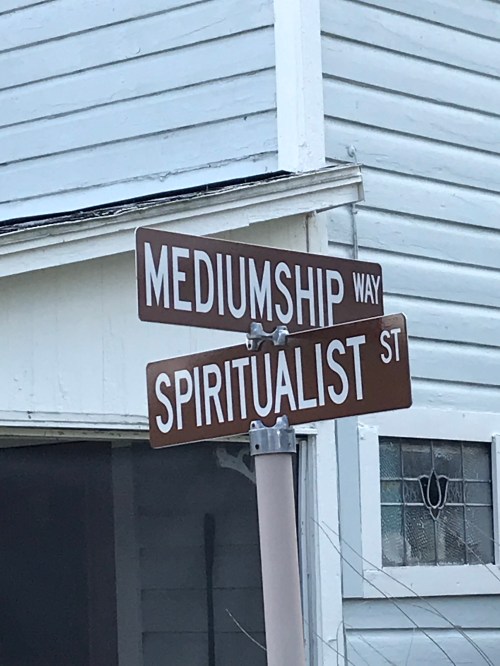 Street signs for "Mediumship Way" and "Spiritualist St" on a building with a white exterior.