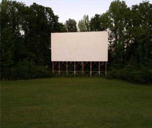 A blank outdoor movie screen stands on wooden supports in a grassy area surrounded by trees.