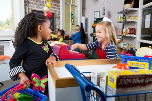 Two children play in a pretend grocery store, one at the register and the other with a shopping basket.