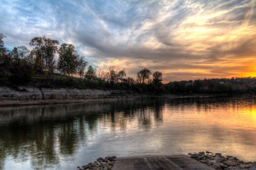 A serene river scene at sunset, with colorful clouds reflecting on the water and trees lining the shore.