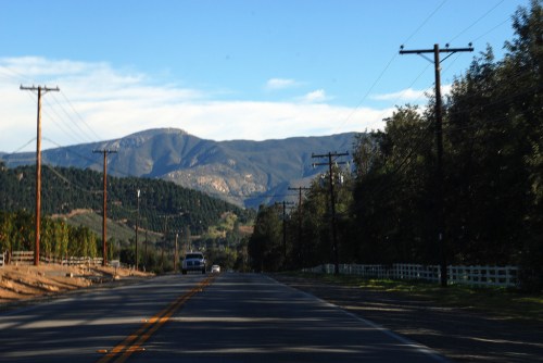 A scenic road lined with power lines, leading towards mountains under a clear blue sky.