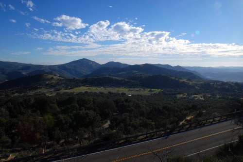 A scenic view of rolling hills and mountains under a blue sky with scattered clouds, alongside a winding road.