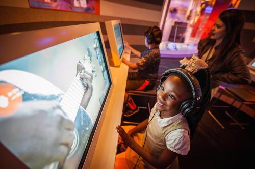 A young girl smiles while wearing headphones at a computer, watching a guitar tutorial, with another child in the background.