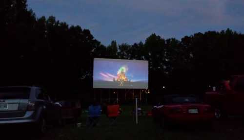 Outdoor movie screen displaying a colorful scene at dusk, surrounded by parked cars and trees.