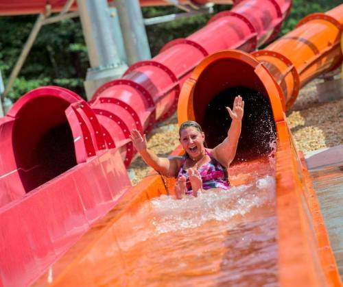 A person joyfully slides down a water slide, splashing into the pool below. Bright orange and red slides are in the background.