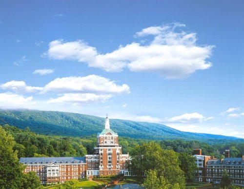 A scenic view of a large building surrounded by trees and mountains under a clear blue sky with fluffy clouds.