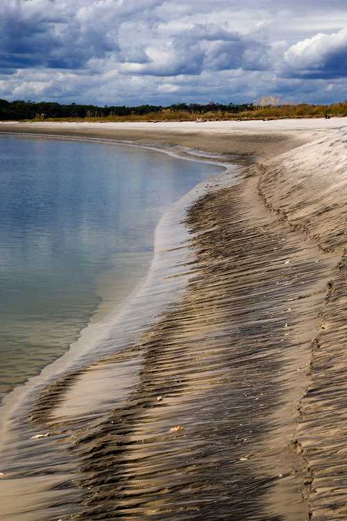 A sandy shoreline curves along a calm water body, with clouds reflecting in the water and greenery in the background.