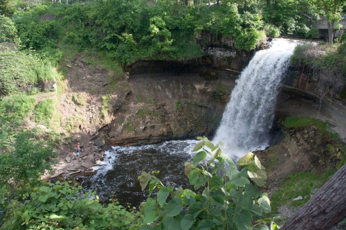A scenic waterfall cascades into a rocky pool, surrounded by lush greenery and trees. Two people stand near the water's edge.