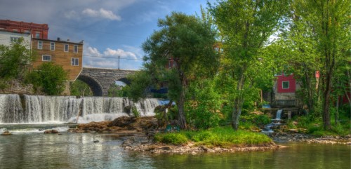 A serene river scene featuring a waterfall, lush greenery, and colorful buildings along the bank under a blue sky.