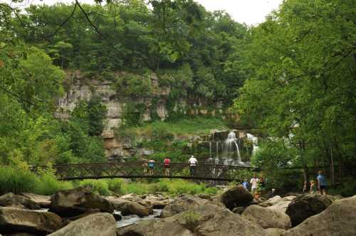 A scenic view of a waterfall surrounded by lush greenery, with people walking on a wooden bridge and exploring the area.