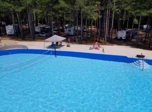 Aerial view of a blue swimming pool surrounded by trees and camping trailers, with people relaxing nearby.