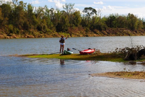 A person stands on a small island by a river, next to green and red kayaks, surrounded by trees and blue sky.