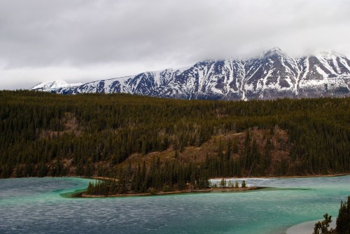 A serene landscape featuring a turquoise lake, dense forests, and snow-capped mountains under a cloudy sky.