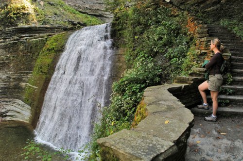 A hiker stands by a waterfall, surrounded by lush greenery and rocky steps in a serene natural setting.