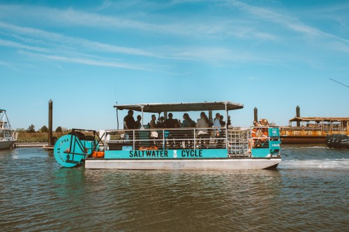 A boat named "Saltwater Cycle" with passengers cruising on a calm waterway under a clear blue sky.