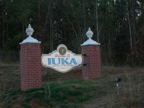 Welcome sign for Iuka, Mississippi, featuring brick pillars and decorative urns, surrounded by trees.