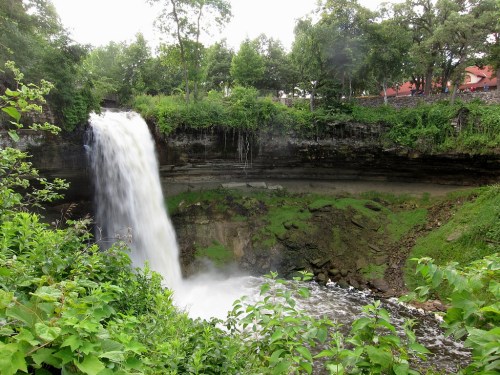 A cascading waterfall surrounded by lush greenery and trees, with a rocky cliff and a hint of a building in the background.