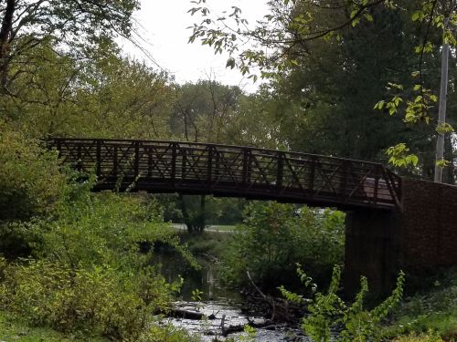 A wooden bridge spans a small creek, surrounded by lush greenery and trees under a clear sky.