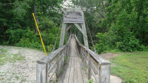 A wooden suspension bridge leads into a lush green forest, with a sign at the entrance.