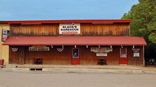 A rustic wooden building with a red roof, featuring signs for Black's Barbecue and open days, surrounded by trees.