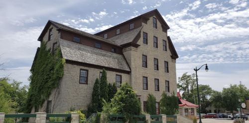 Historic stone building with a steep roof, surrounded by greenery and under a partly cloudy sky.