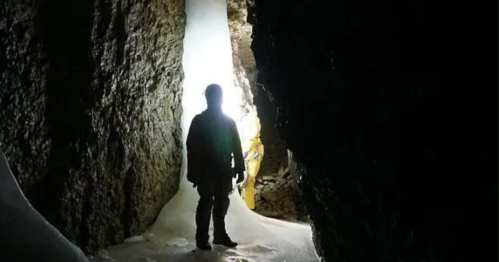 A silhouetted figure stands in a dark cave, illuminated by light from an icy opening ahead.