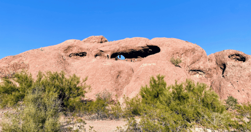 A large reddish rock formation with a natural arch, surrounded by sparse vegetation under a clear blue sky.