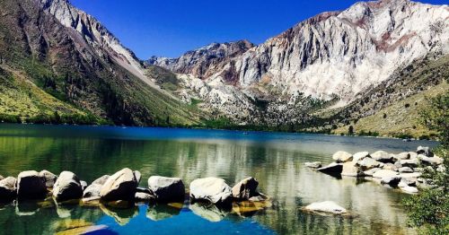 Scenic lake surrounded by mountains, with clear blue water reflecting the landscape and rocky shoreline.
