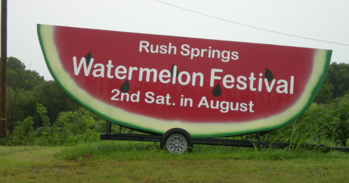 A large sign for the Rush Springs Watermelon Festival, featuring a watermelon design and details about the event.