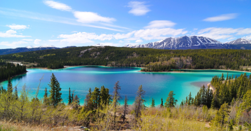 A scenic view of a turquoise lake surrounded by green trees and mountains under a blue sky with scattered clouds.