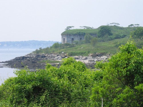 A grassy hill with a stone structure overlooks a calm body of water, surrounded by lush greenery and rocky shoreline.