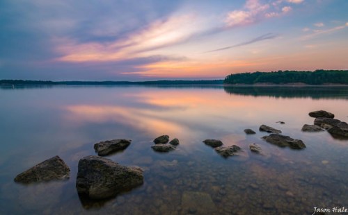 A serene lake at sunset, with calm waters reflecting colorful clouds and rocky shoreline in the foreground.