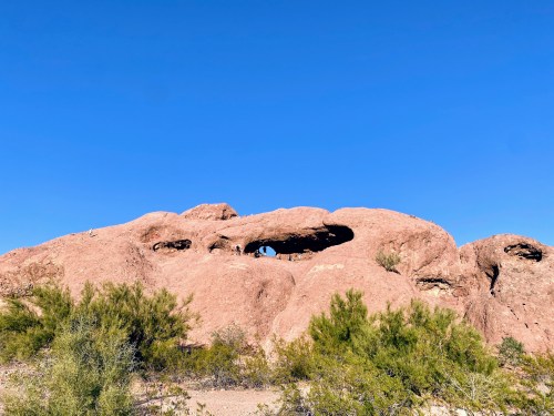 A large reddish rock formation with a prominent arch, set against a clear blue sky and surrounded by sparse vegetation.