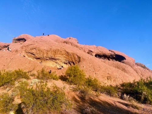 A rocky, reddish-brown hillside with people exploring caves under a clear blue sky. Green shrubs are in the foreground.