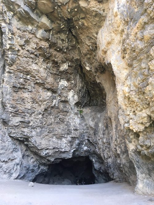A large rocky cave entrance with rugged stone walls and a dark interior, surrounded by natural terrain.