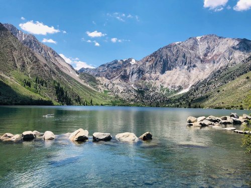 A serene lake surrounded by mountains, with clear blue water and a rocky shoreline under a bright sky.