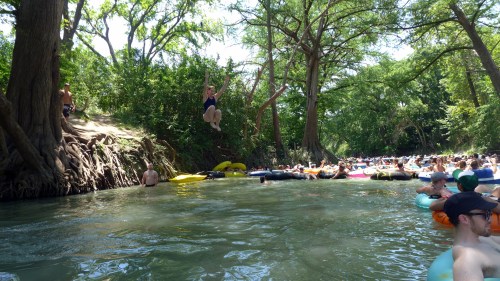A lively river scene with people floating on tubes and someone jumping from a tree into the water. Lush greenery surrounds the area.