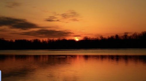 A serene sunset over a calm lake, with orange and yellow hues reflecting on the water and silhouettes of trees in the background.
