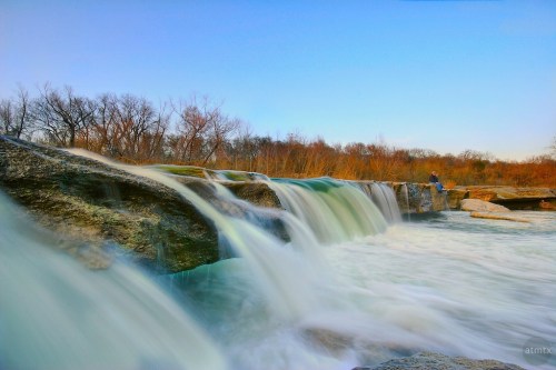 A serene waterfall cascades over rocks, surrounded by trees under a clear blue sky. A person sits nearby, enjoying the view.