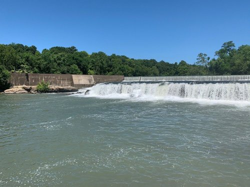 A serene waterfall cascades over a dam into a calm river, surrounded by lush green trees under a clear blue sky.