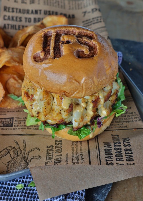 A gourmet chicken sandwich with lettuce and cheese, served on a toasted bun with "JFS" embossed on top, alongside potato chips.