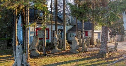A quirky, weathered house surrounded by tall trees and unique stone sculptures in a serene outdoor setting.