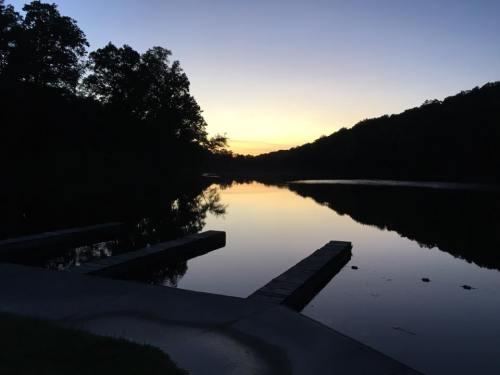 Silhouette of trees and a calm lake at sunset, with reflections on the water and a serene atmosphere.