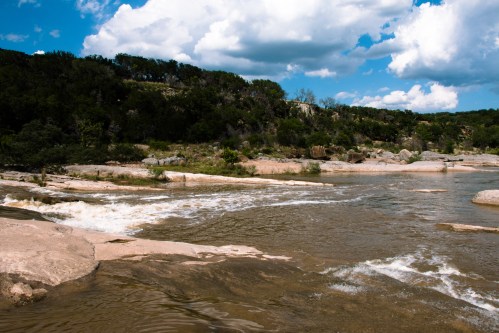 A serene river flows over rocky terrain, surrounded by lush greenery and a blue sky with fluffy clouds.